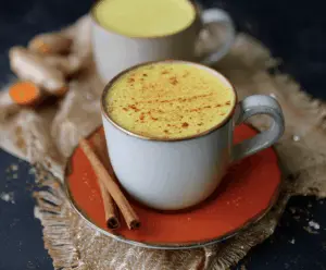 A cozy cup of Grounding Autumn Ginger Turmeric Latte topped with cinnamon and ginger, served in a rustic mug with fresh ginger and turmeric root, perfect for a warming fall beverage.
