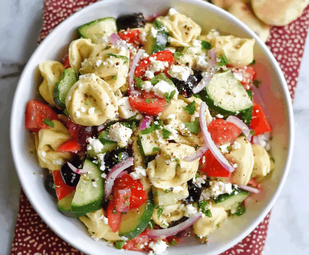Colorful Greek Tortellini Pasta Salad with cherry tomatoes, cucumbers, olives, feta cheese, and fresh herbs in a bowl, perfect for summer picnics and parties.
