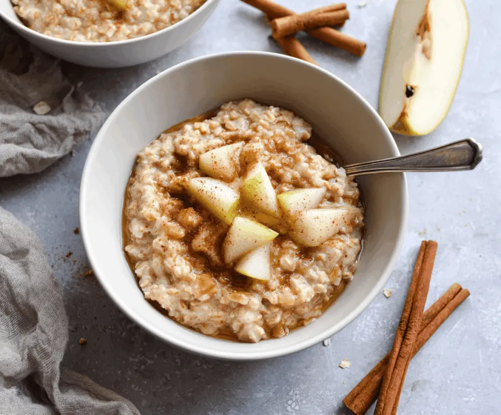 A bowl of warm spiced pear oatmeal topped with cinnamon and fresh pear slices, served with a spoon on a rustic wooden table.